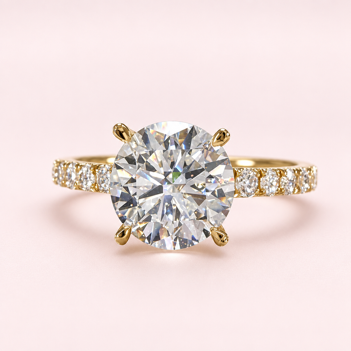 Front view of a round diamond engagement ring with yellow-gold prongs and a diamond-set band on a pale pink background.
