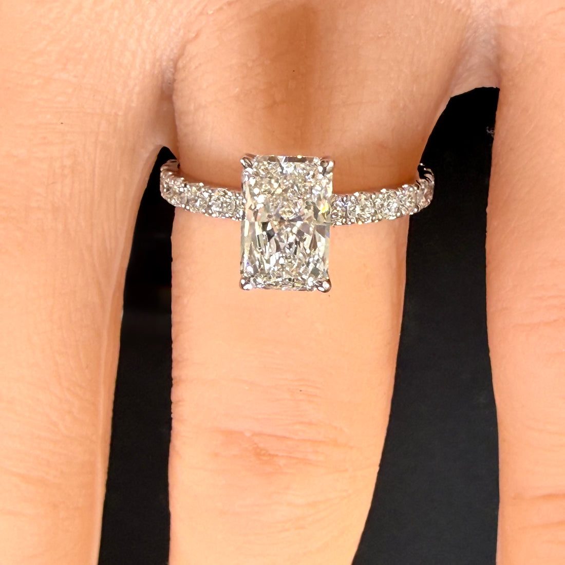 Close-up of a radiant-cut diamond ring with a white-gold pavé band worn on a finger.
