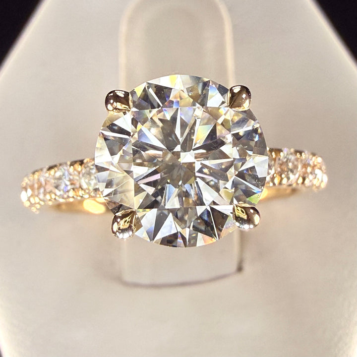 Front close-up of a round diamond engagement ring with four yellow-gold prongs and a pavé band.