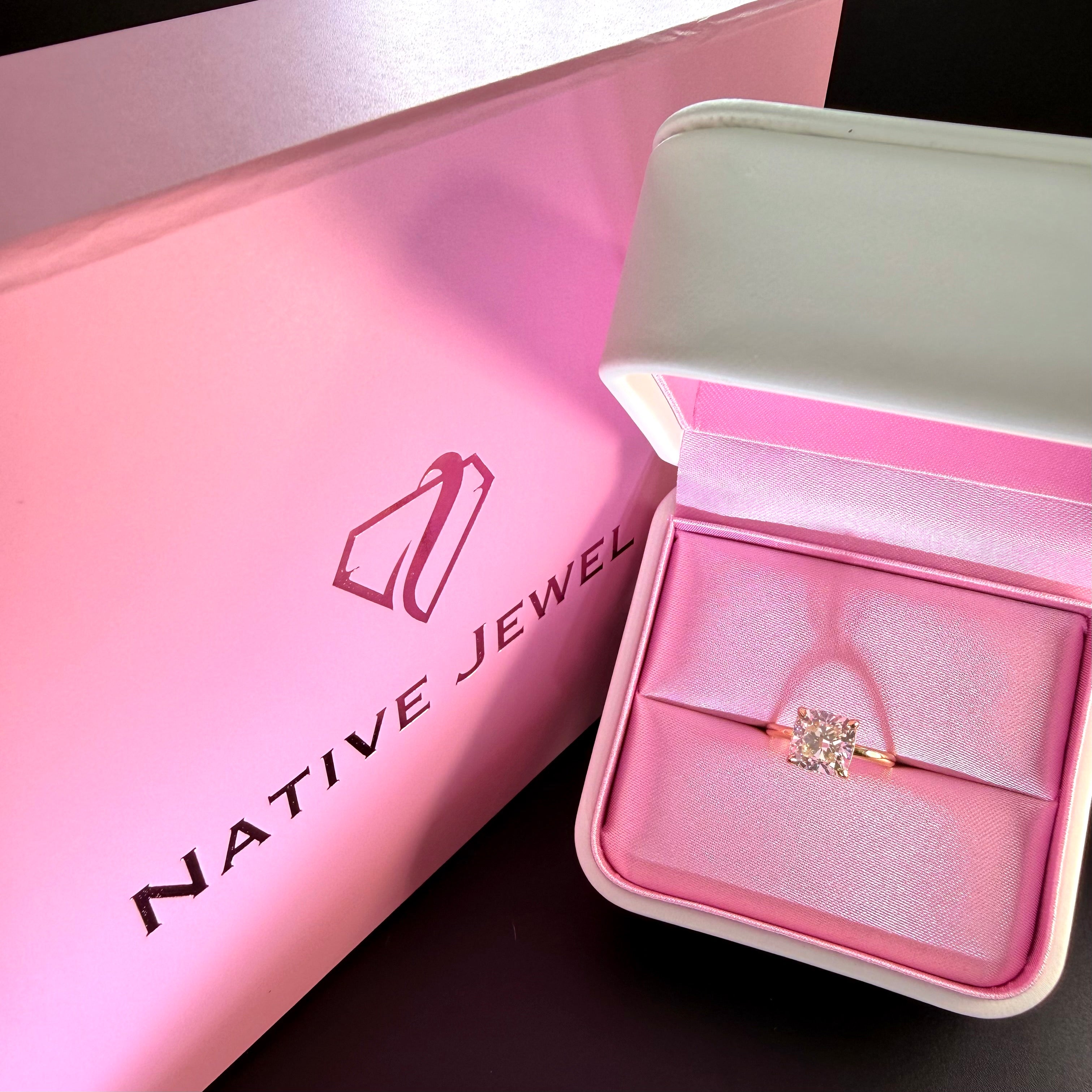 Diamond solitaire with a yellow-gold band in an open pink-lined presentation box beside a Native Jewel bag.
