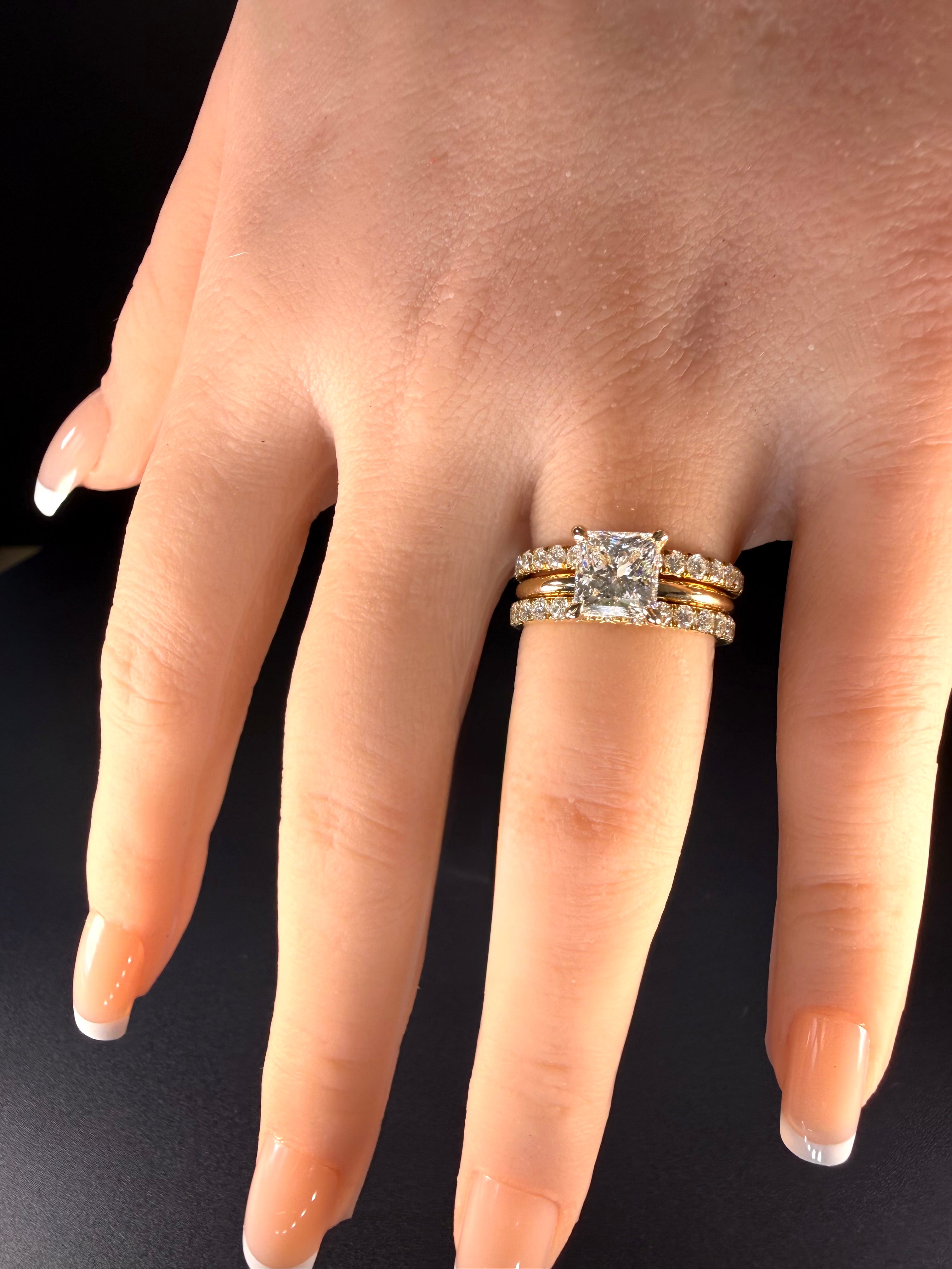 Princess-cut diamond solitaire in yellow gold worn between two diamond-set bands on a hand.