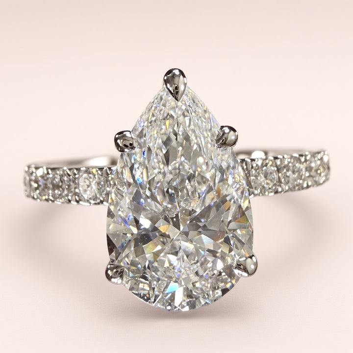 Front close-up of a pear-shaped diamond engagement ring with a white-gold pavé band on pale pink.