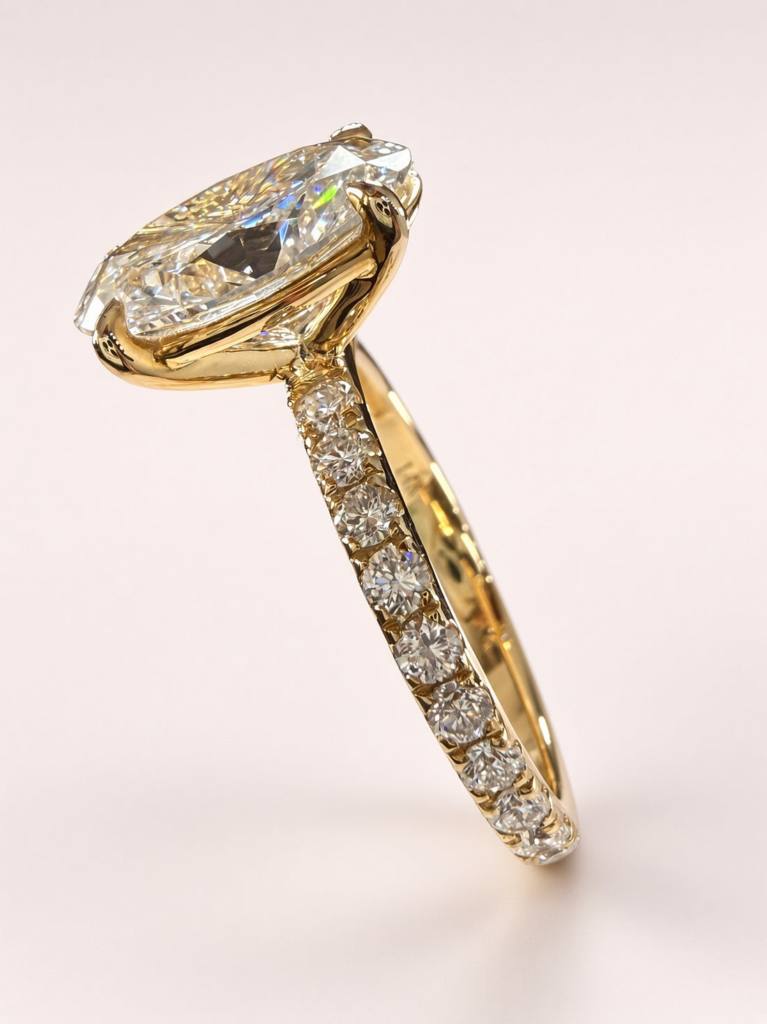 Angled view of an oval diamond pavé ring in yellow gold on a pale pink background.