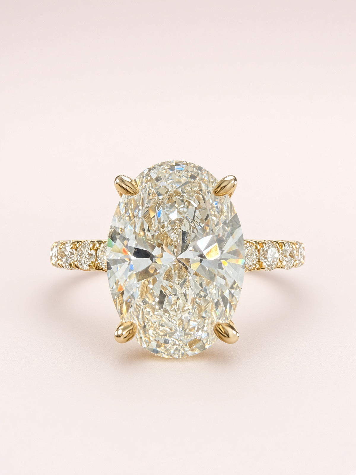 An oval diamond engagement ring featuring a large center stone in a yellow gold four-prong setting, with a diamond-accented band, set against a soft light pink background.