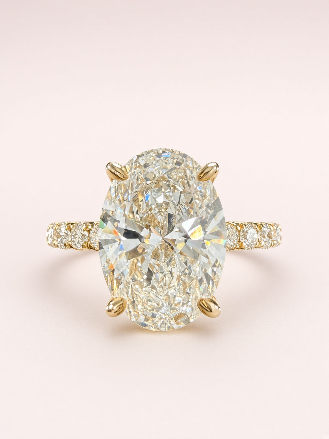 An oval diamond engagement ring featuring a large center stone in a yellow gold four-prong setting, with a diamond-accented band, set against a soft light pink background.