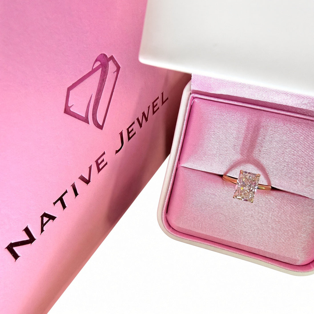 Radiant-cut diamond solitaire in a pink-lined presentation box beside a pink Native Jewel shopping bag.