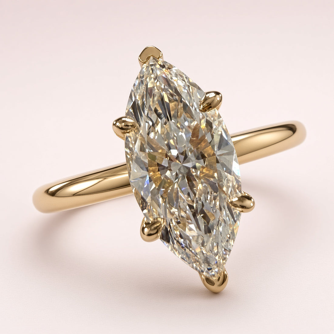 Angled front close-up of a marquise diamond solitaire with yellow-gold tip guards and a plain band on pale pink.