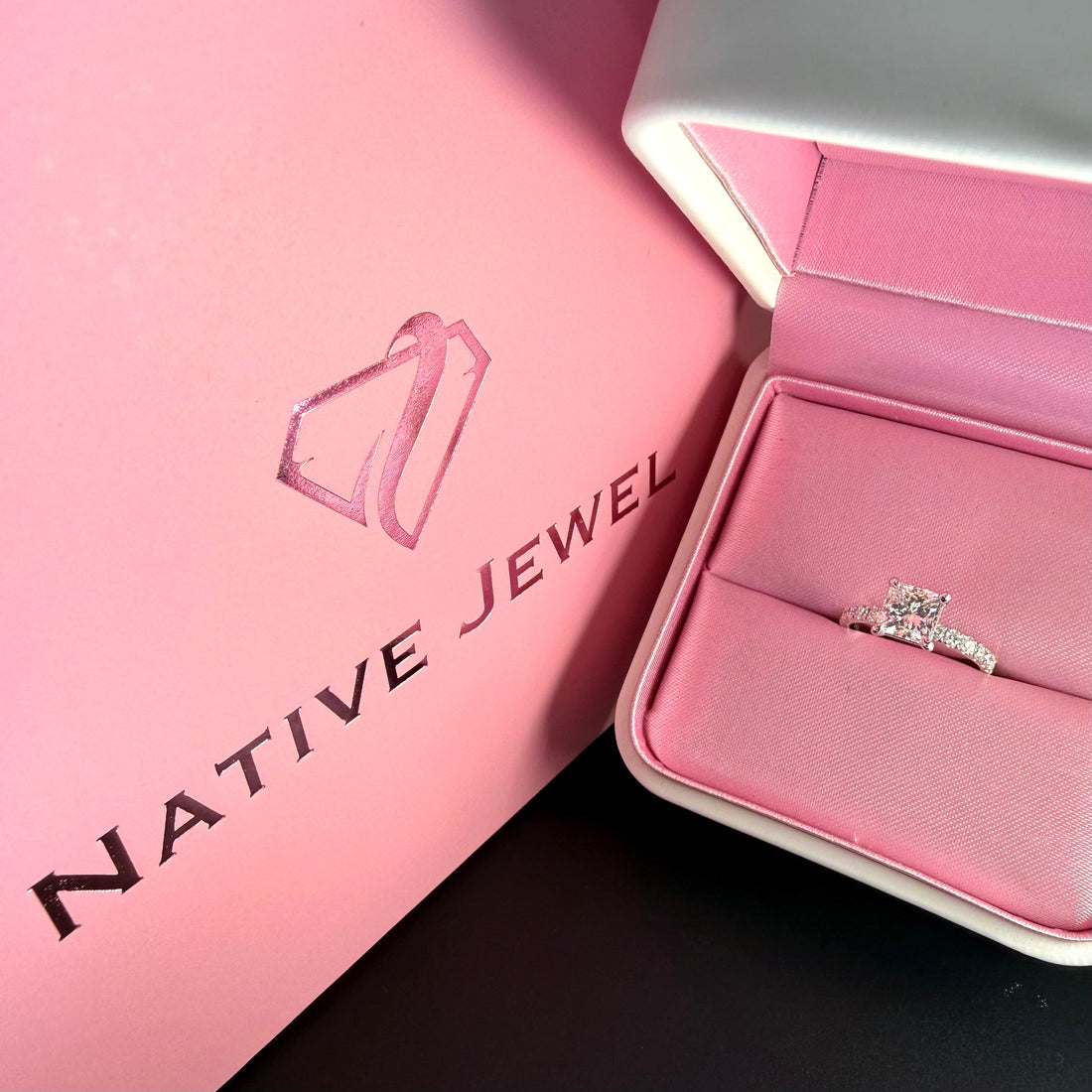 Radiant-cut diamond ring with a diamond-set band in a pink ring box beside Native Jewel packaging.