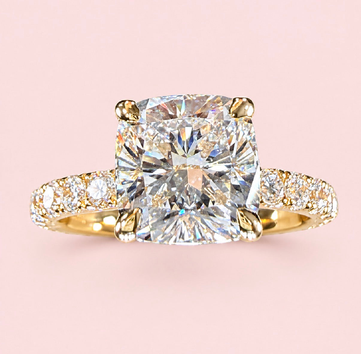 Front view of a cushion-cut diamond ring in yellow gold with a diamond-set band on a pink background.