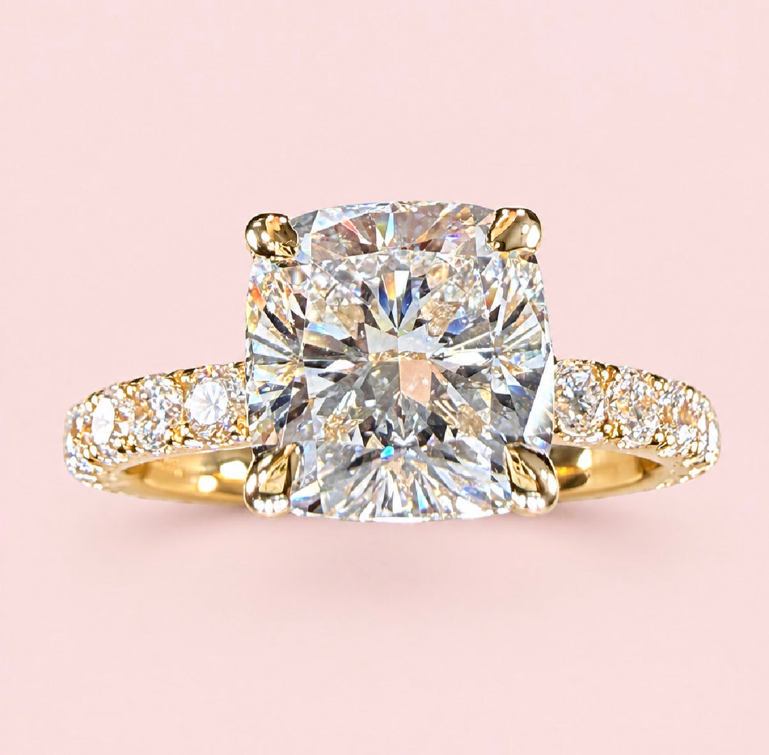 Front view of a cushion-cut diamond ring in yellow gold with a diamond-set band on a pink background.