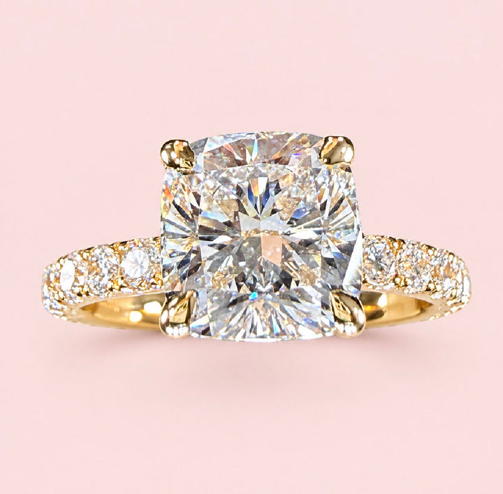 Front view of a cushion-cut diamond ring in yellow gold with a diamond-set band on a pink background.
