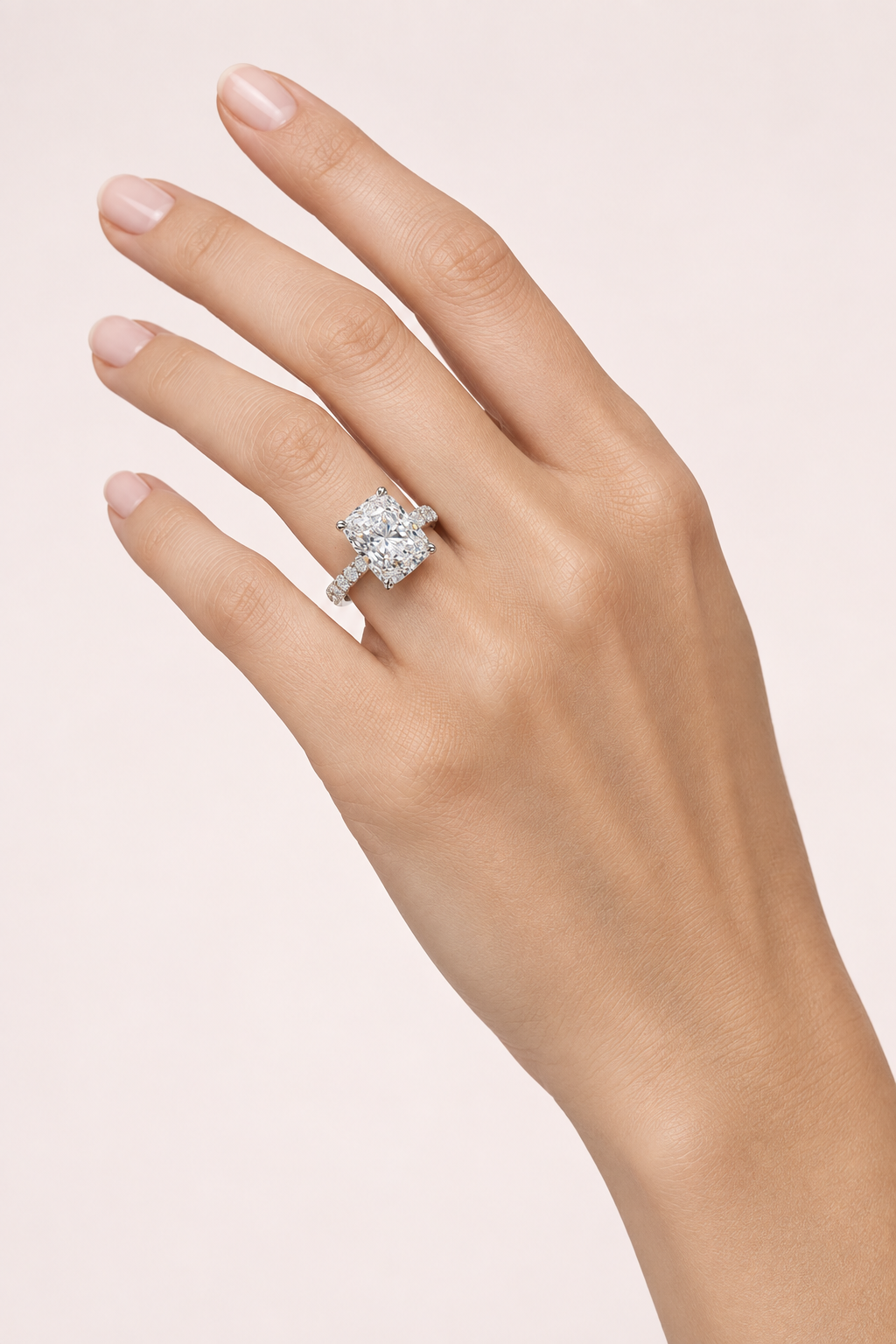 Cushion-cut diamond engagement ring with a white-gold diamond-set band worn on a hand against pale pink.