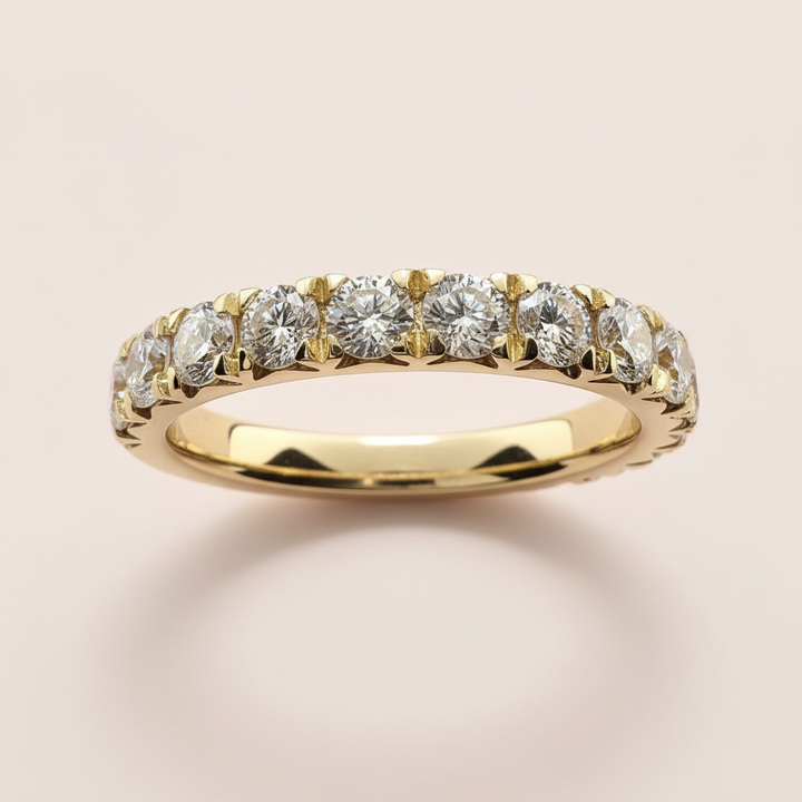 A yellow gold wedding band featuring 14k yellow gold ring,  row of round-cut clear gemstones in a prong setting, shown on a soft, light beige background.