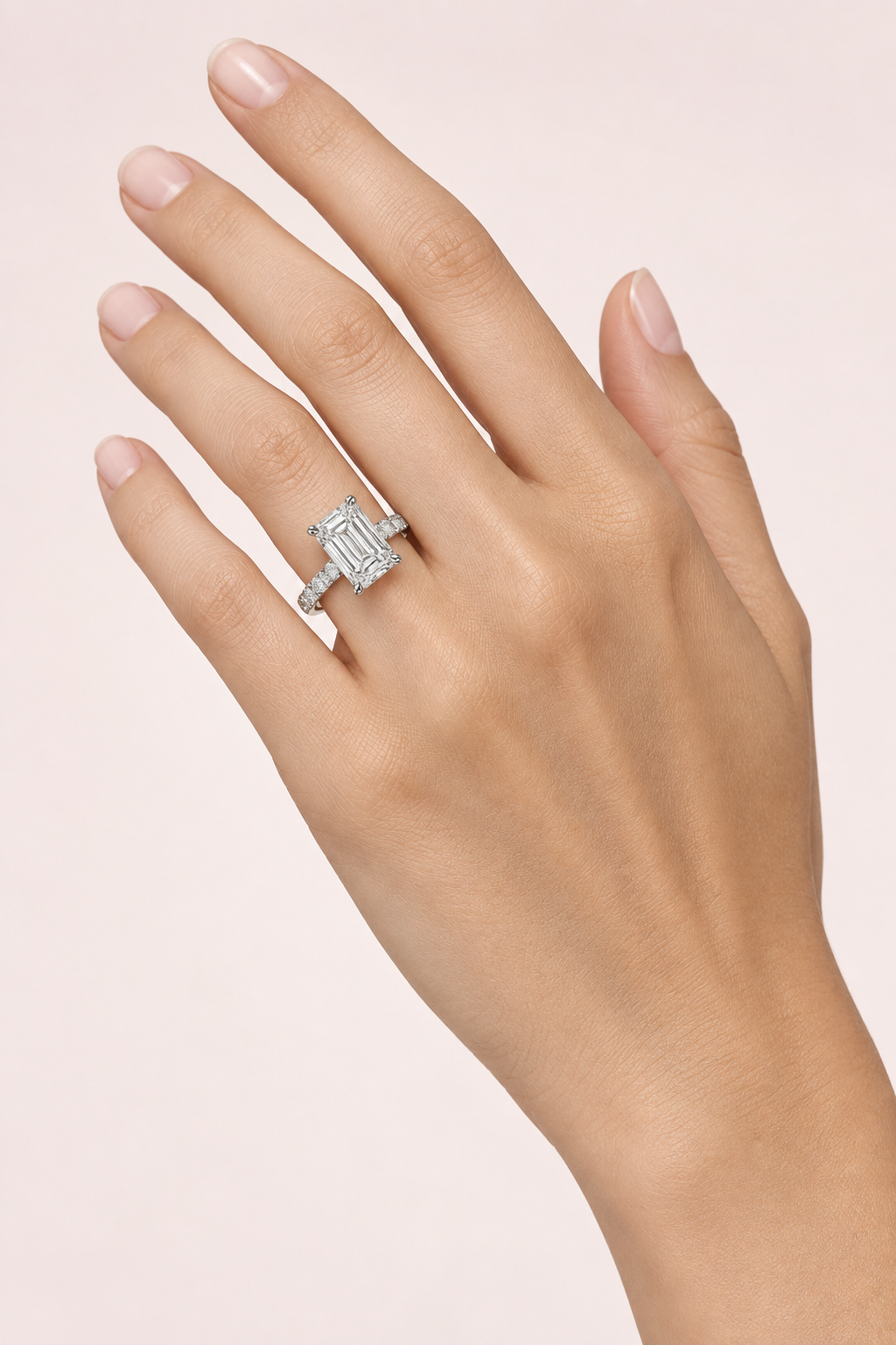 Emerald-cut diamond engagement ring with a white-gold diamond-set band worn on a hand against pale pink.