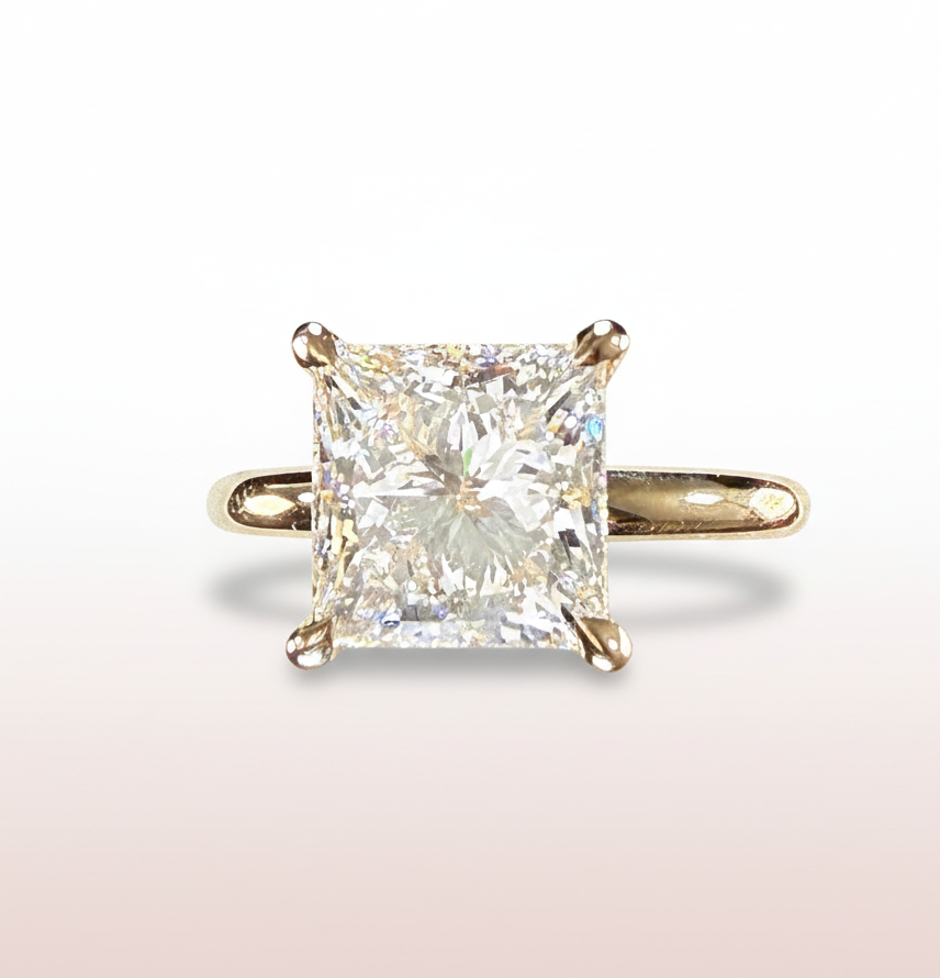 A princess cut diamond engagement ring in a yellow gold four-prong setting, centered on a clean white-to-pale-pink gradient background.