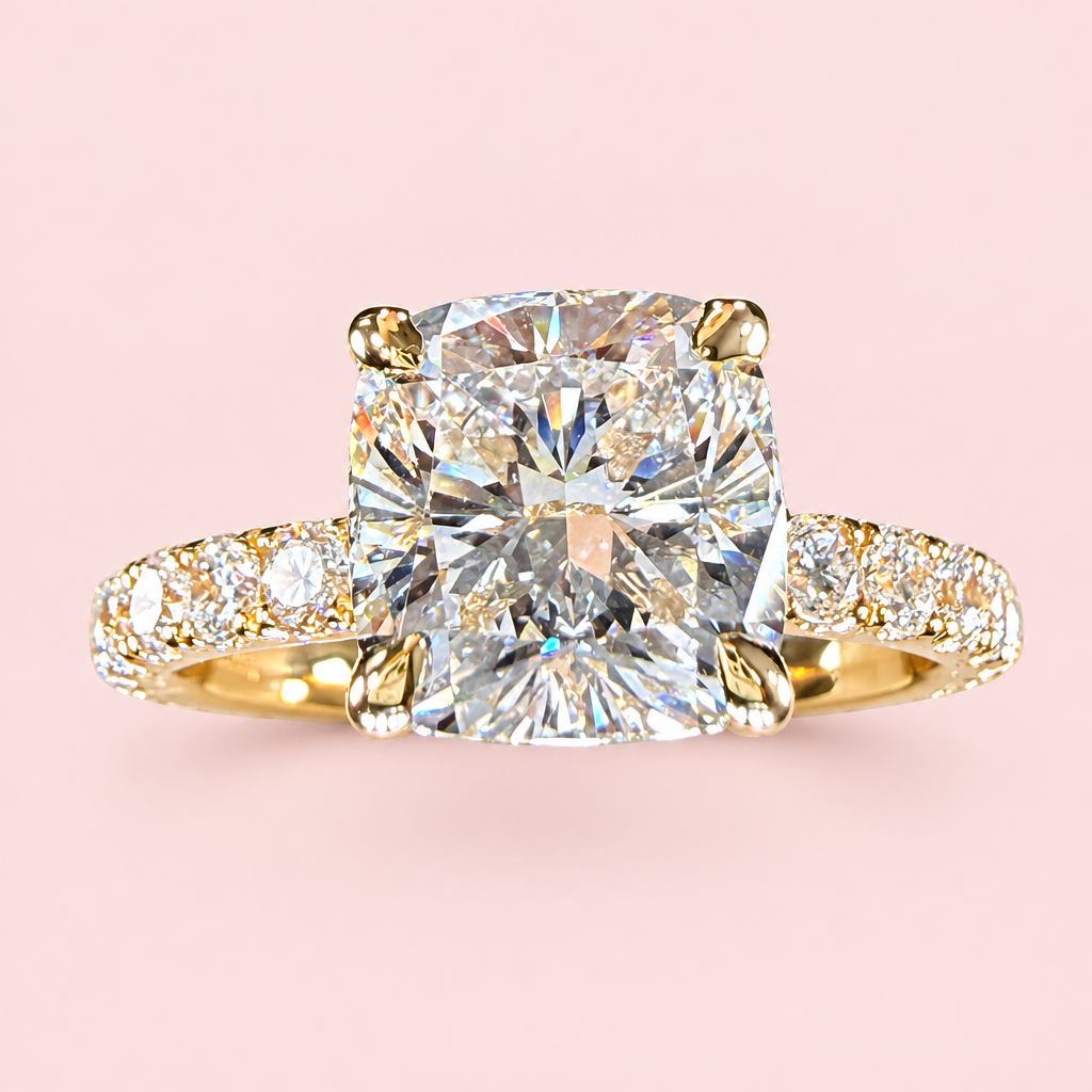A yellow gold engagement ring featuring a large cushion-cut diamond center stone and a pavé-set diamond band, shown against a solid pale pink background.